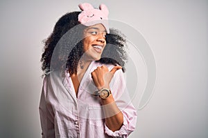 Young african american woman with afro hair wearing sleeping eye mask and pink pajama smiling with happy face looking and pointing