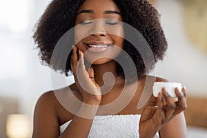 Young African American woman applying hydrating cream on her face, smiling with closed eyes, indoors