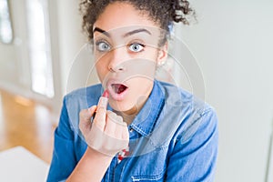 Young african american woman applying red lipstick on lips scared in shock with a surprise face, afraid and excited with fear