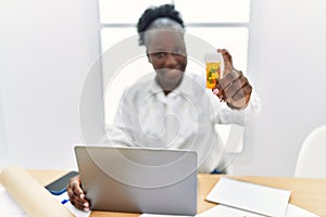 Young african american woman architect using laptop holding pills at architecture studio