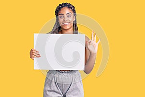 Young african american woman with braids holding blank empty banner doing ok sign with fingers, smiling friendly gesturing