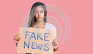 Young african american woman with braids holding fake news banner thinking attitude and sober expression looking self confident