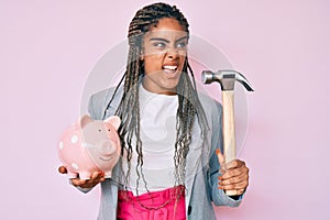 Young african american woman with braids holding piggy bank and hammer angry and mad screaming frustrated and furious, shouting