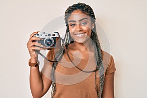 Young african american woman with braids holding vintage camera looking positive and happy standing and smiling with a confident