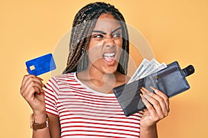 Young african american woman with braids holding wallet with dollars and credit card angry and mad screaming frustrated and