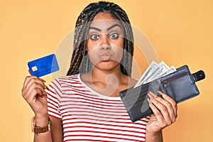 Young african american woman with braids holding wallet with dollars and credit card puffing cheeks with funny face