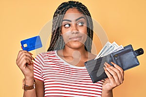 Young african american woman with braids holding wallet with dollars and credit card smiling looking to the side and staring away