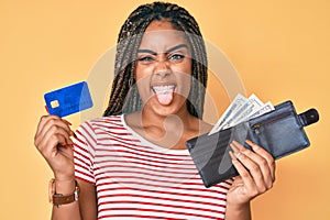 Young african american woman with braids holding wallet with dollars and credit card sticking tongue out happy with funny