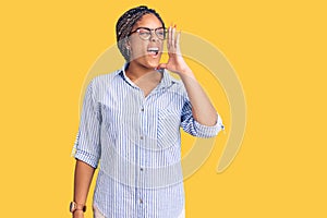 Young african american woman with braids wearing casual clothes and glasses shouting and screaming loud to side with hand on mouth