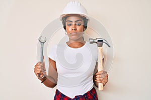 Young african american woman with braids wearing hardhat holding hammer and wrench relaxed with serious expression on face