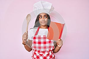 Young african american woman with braids wearing professional baker apron reading cooking recipe book smiling looking to the side