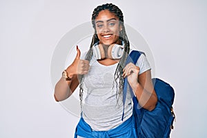 Young african american woman with braids wearing student backpack and headphones smiling happy and positive, thumb up doing