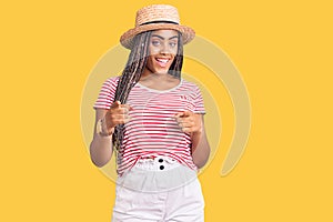 Young african american woman with braids wearing summer hat pointing fingers to camera with happy and funny face