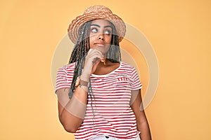 Young african american woman with braids wearing summer hat serious face thinking about question with hand on chin, thoughtful