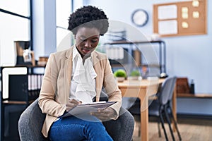 Young african american woman business worker writing on paperwork sitting on chair at office