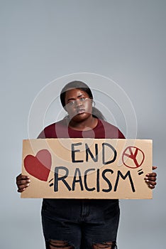 Young african american woman in casual clothes holding End Racism banner in front of her, posing isolated over gray