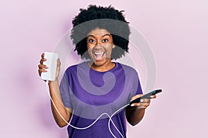 Young african american woman charging smartphone using portable battery celebrating crazy and amazed for success with open eyes