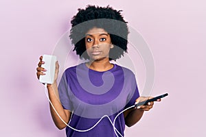 Young african american woman charging smartphone using portable battery relaxed with serious expression on face