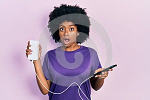 Young african american woman charging smartphone using portable battery in shock face, looking skeptical and sarcastic, surprised