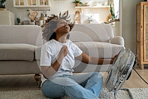 Young African American woman cooling down by ventilator at home, feeling unwell during heatwave