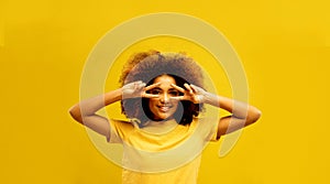 Young african american woman covering eye with victory v-sign gesture isolated on yellow color background.