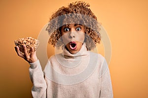 Young african american woman with curly hair holding bowl with healthy peanuts scared in shock with a surprise face, afraid and