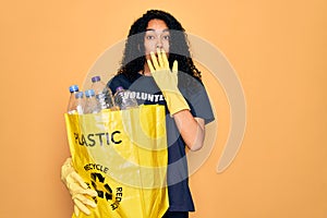 Young african american woman doing volunteering recycling holding bag with plastic bottles cover mouth with hand shocked with