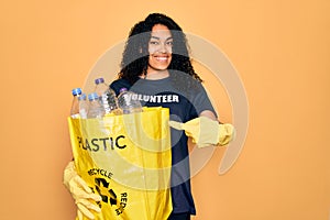 Young african american woman doing volunteering recycling holding bag with plastic bottles very happy pointing with hand and