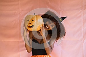 Young african american woman dressed as a witch for halloween party and holding a pumpkin in her hands. She wears a witch`s hat