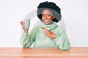 Young african american woman drinking cup of coffee smiling happy pointing with hand and finger