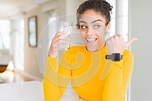 Young african american woman drinking a glass of fresh water pointing and showing with thumb up to the side with happy face