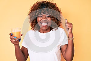 Young african american woman drinking glass of orange juice screaming proud, celebrating victory and success very excited with