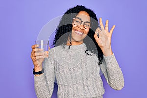 Young african american woman drinking glass of water over isolated purple background doing ok sign with fingers, excellent symbol