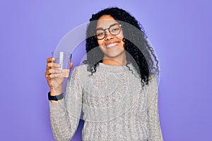 Young african american woman drinking glass of water over isolated purple background with a happy face standing and smiling with a