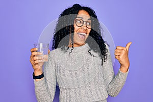 Young african american woman drinking glass of water over isolated purple background pointing and showing with thumb up to the