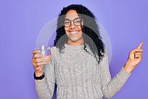 Young african american woman drinking glass of water over isolated purple background very happy pointing with hand and finger to