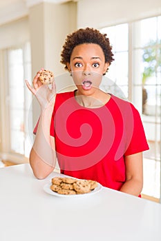 Young african american woman eating chocolate chips cookies scared in shock with a surprise face, afraid and excited with fear