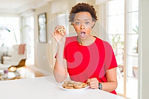 Young african american woman eating chocolate chips cookies scared in shock with a surprise face, afraid and excited with fear