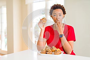 Young african american woman eating chocolate chips muffins cover mouth with hand shocked with shame for mistake, expression of