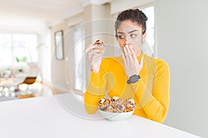 Young african american woman eating chocolate chips muffins cover mouth with hand shocked with shame for mistake, expression of