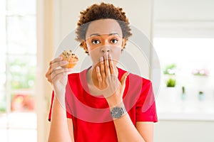 Young african american woman eating chocolate chips muffins cover mouth with hand shocked with shame for mistake, expression of