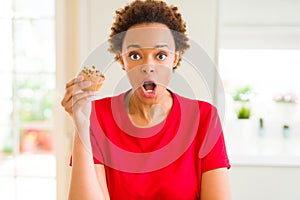 Young african american woman eating chocolate chips muffins scared in shock with a surprise face, afraid and excited with fear