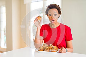 Young african american woman eating chocolate chips muffins scared in shock with a surprise face, afraid and excited with fear