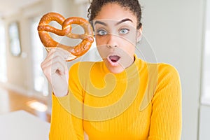Young african american woman eating German salty pastry pretzel scared in shock with a surprise face, afraid and excited with fear