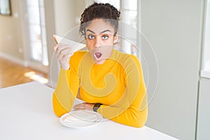 Young african american woman eating a sandwich as healthy snack scared in shock with a surprise face, afraid and excited with fear