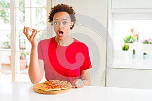 Young african american woman eating tasty peperoni pizza scared in shock with a surprise face, afraid and excited with fear