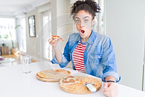 Young african american woman eating two homemade cheese pizzas scared in shock with a surprise face, afraid and excited with fear