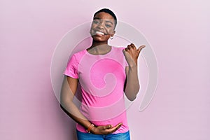 Young african american woman expecting a baby, touching pregnant belly pointing to the back behind with hand and thumbs up,