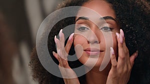 Young African American woman girl touching pretty face applying cream do make up looking in mirror perfect skin care