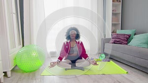 Young african american woman hippy in colourful sportswear with curly afro hairs sits in lotus pose and meditations